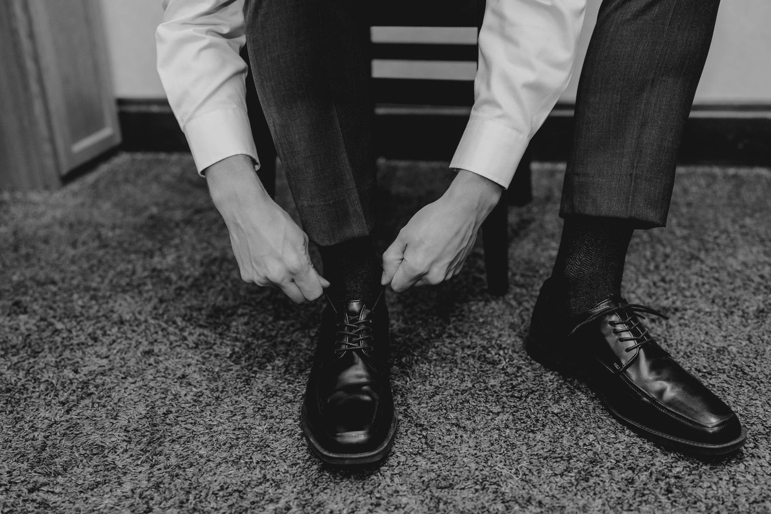 Man in dress shirt and gray trousers tying black oxford shoes, black and white wedding preparation photo.