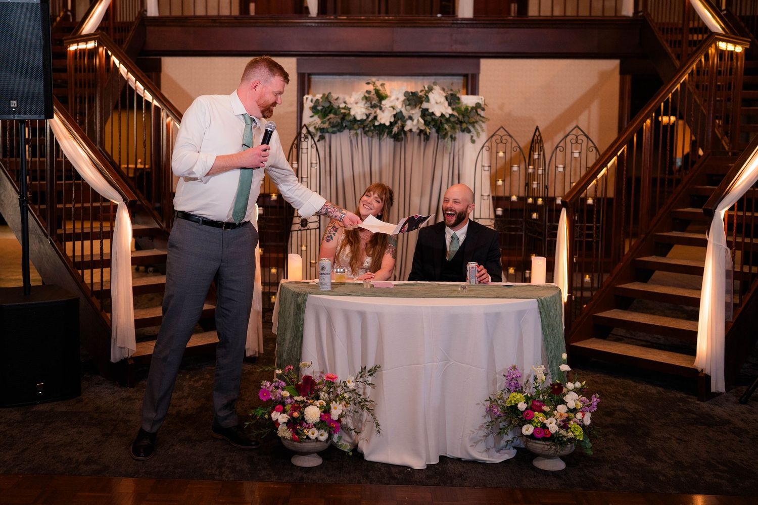 Best man delivers toast to bride and groom at elegant wedding reception with floral arrangements.