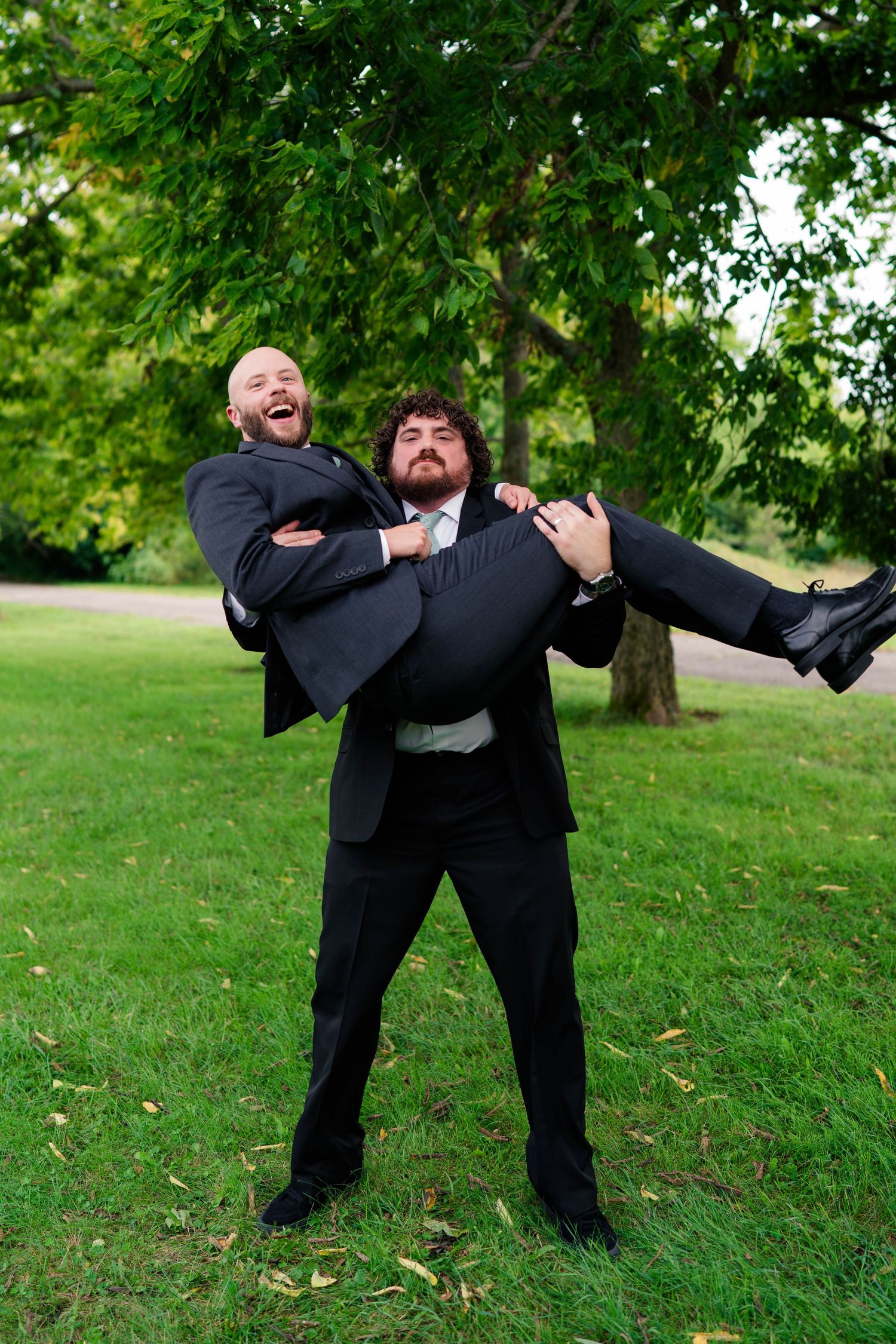 Groomsman in black suit playfully carries another man in arms outdoors at wedding celebration.