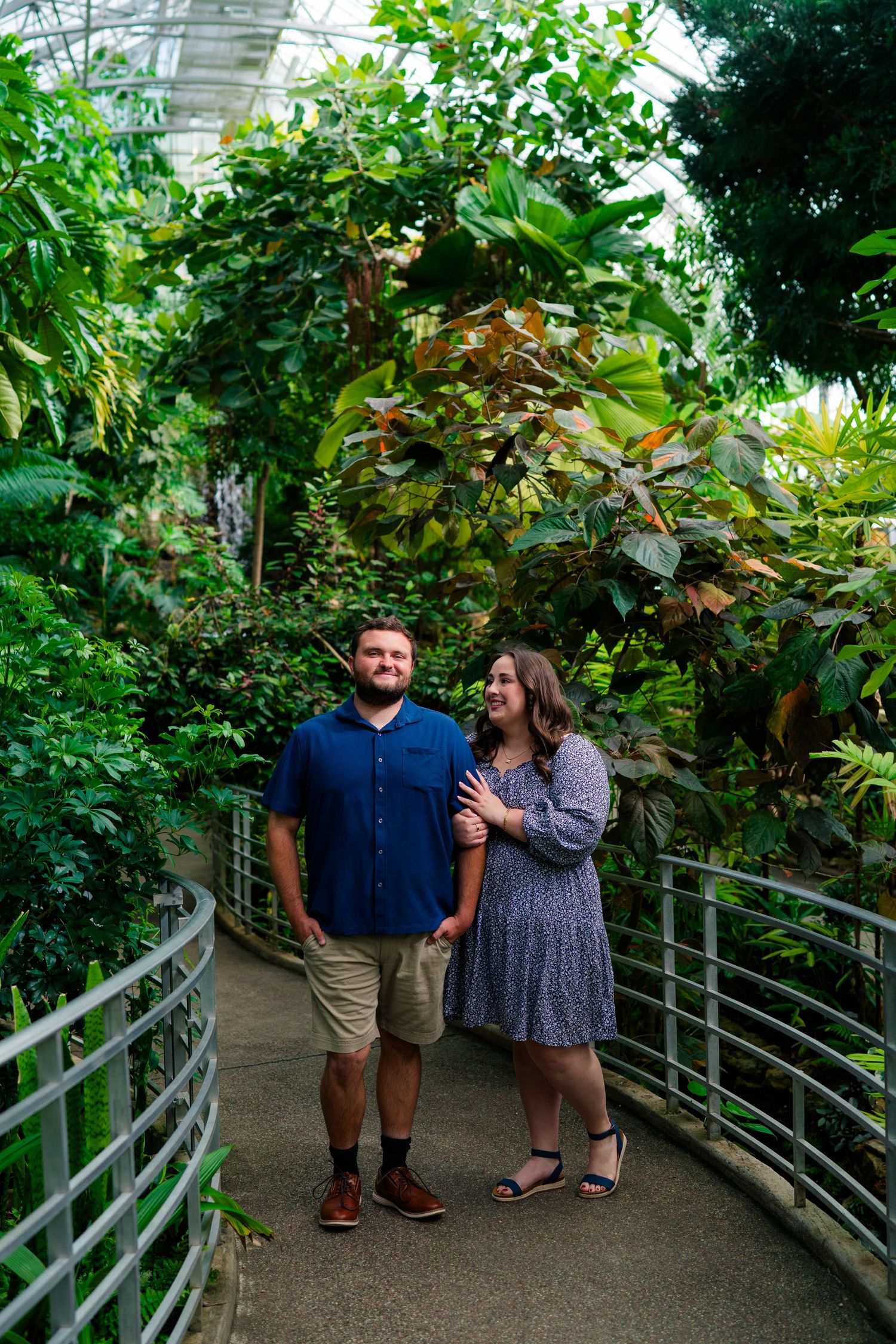 Happy couple walks hand in hand along a path surrounded by vibrant tropical plants in a greenhouse.