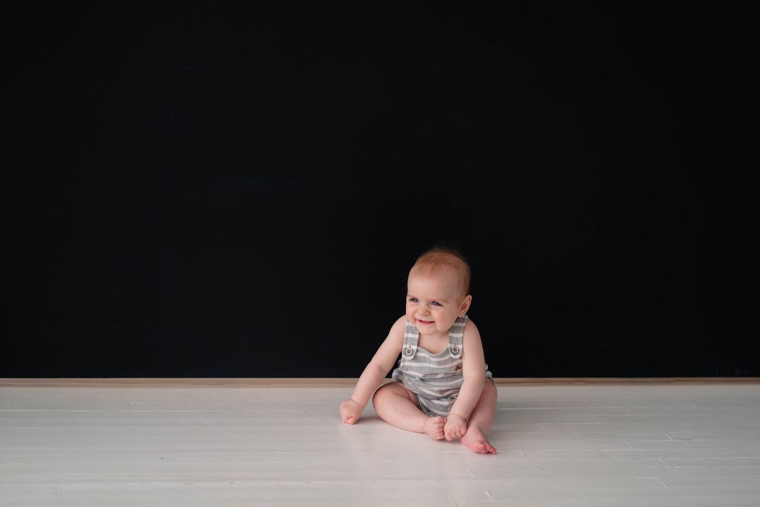 Baby in gray romper sitting on white floor against dramatic dark black background.