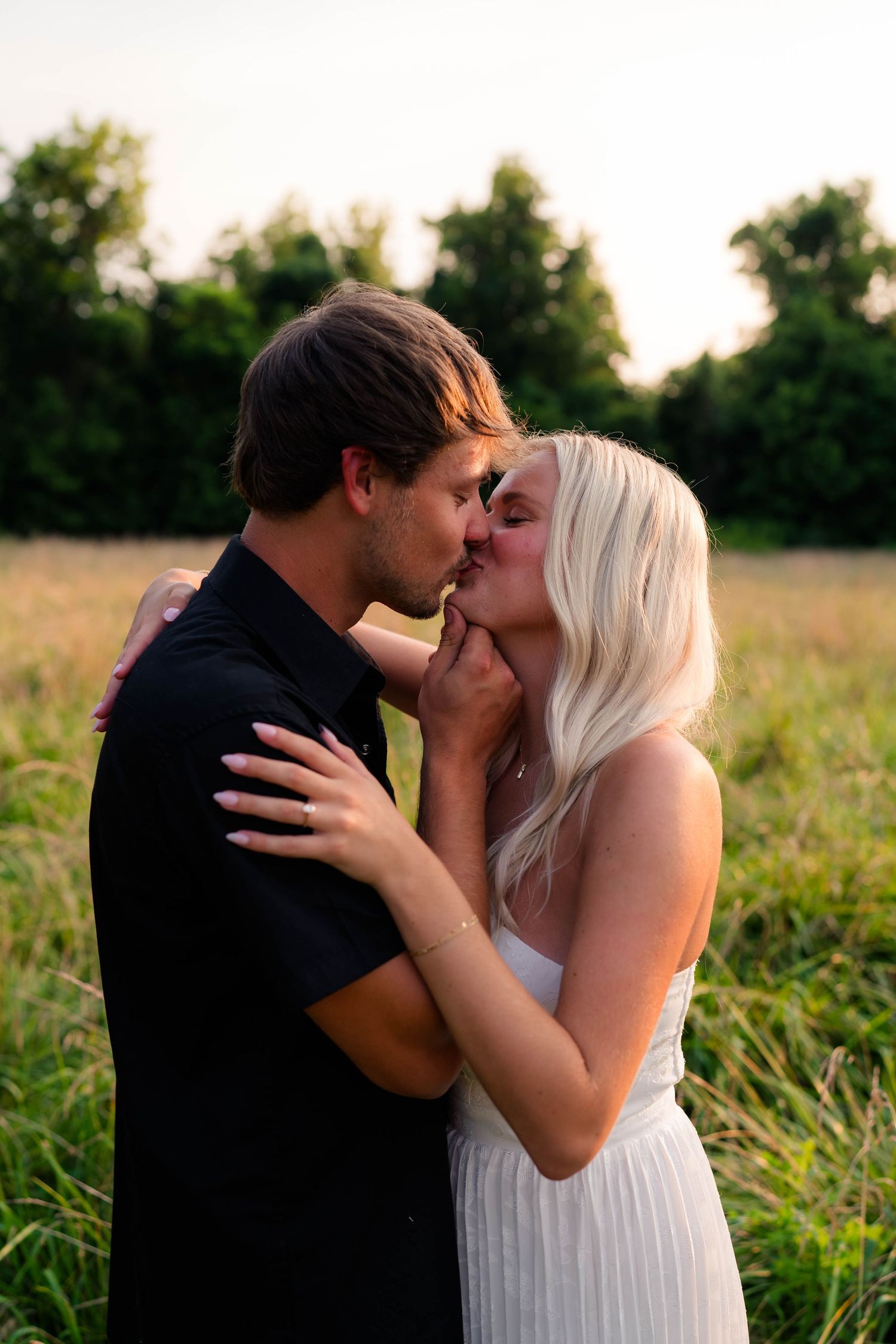 Couple sharing a romantic kiss in a golden field at sunset, woman in white dress with blonde hair.