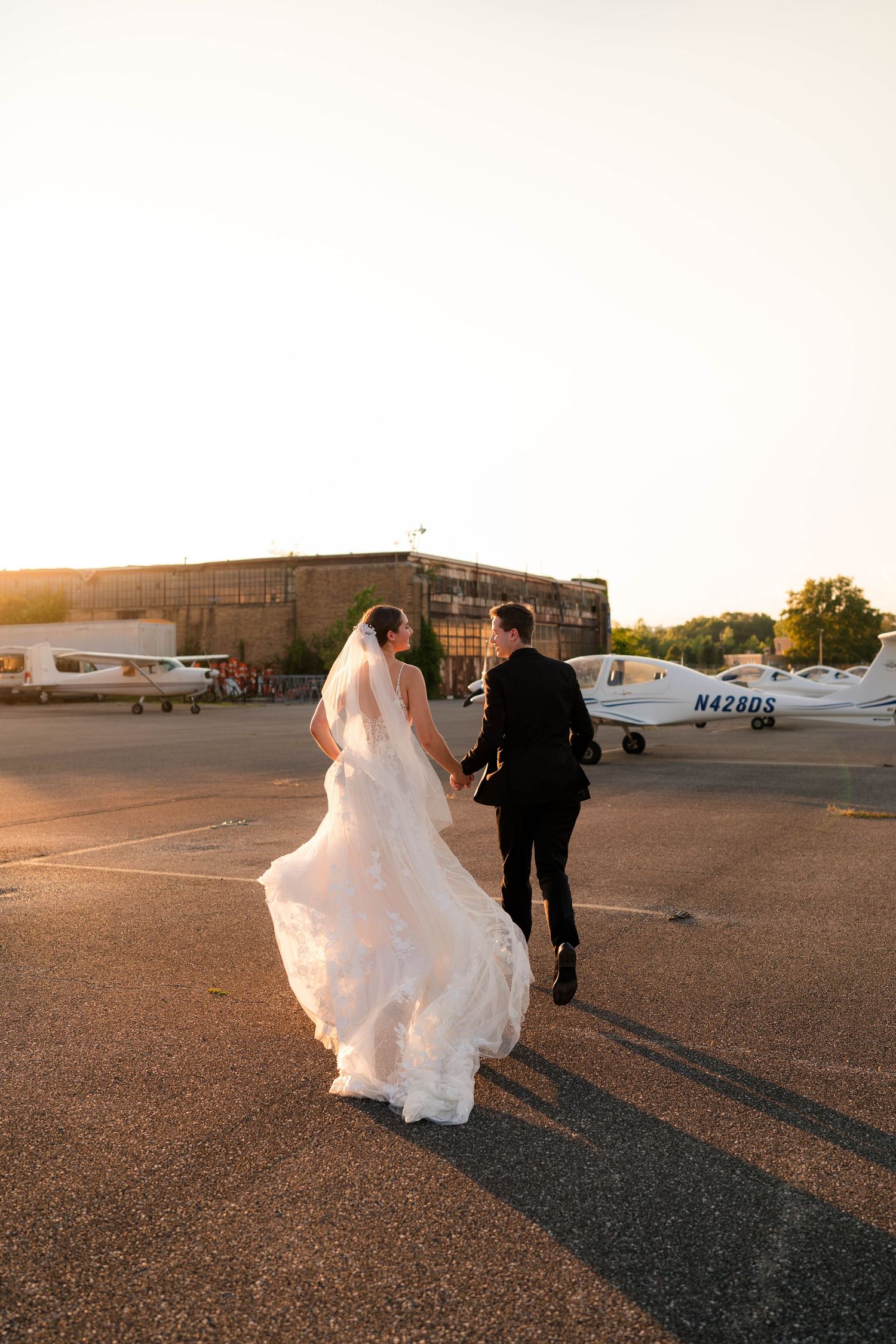 Bride and groom walk hand-in-hand across a sunlit airport tarmac, wedding dress flowing in the breeze.