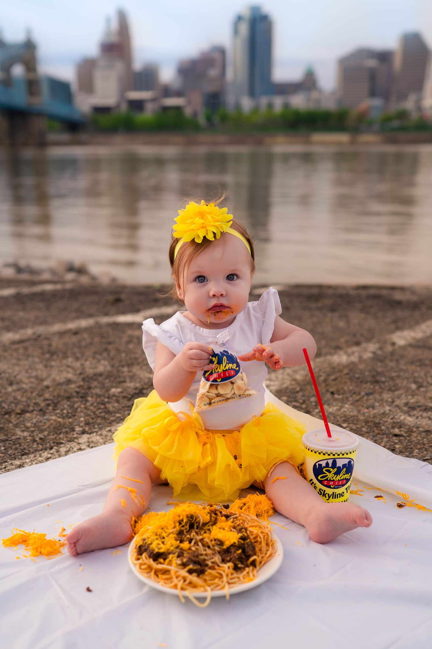 Baby girl in yellow tutu eating Skyline Chili spaghetti on riverbank with Cincinnati skyline backdrop.
