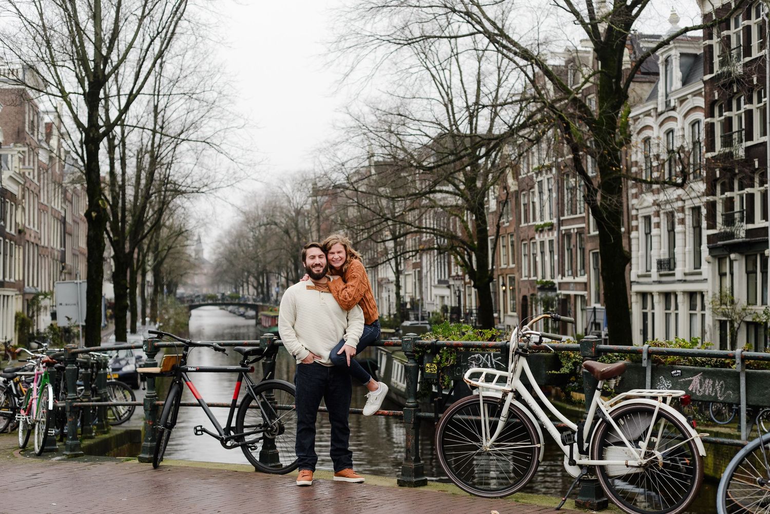Couple posing together on Amsterdam canal bridge surrounded by bicycles and historic buildings.