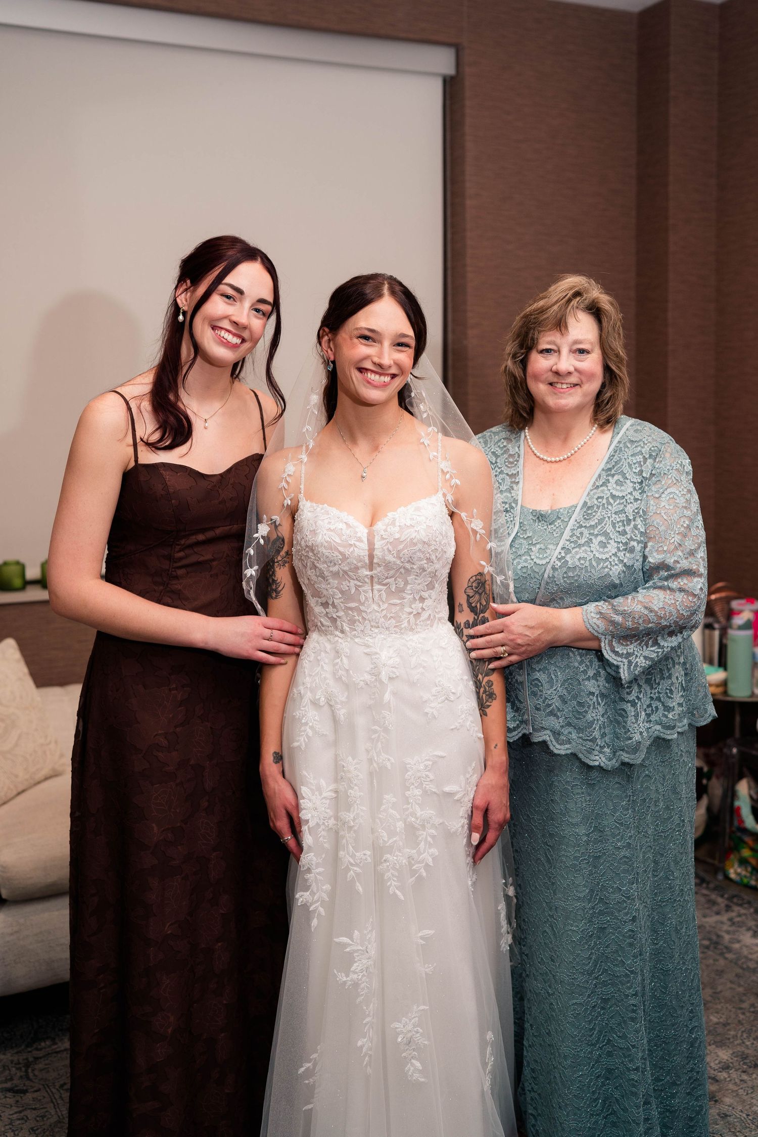 Bride in white lace gown with veil posing with bridesmaid and mother in teal lace dress.
