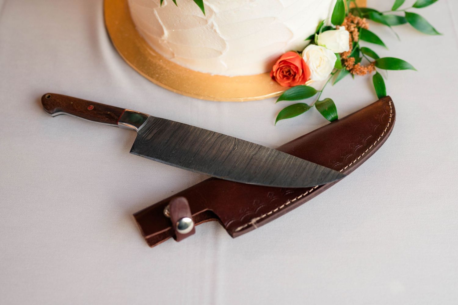 Custom Damascus steel cake knife with leather sheath displayed beside a white two-tier wedding cake.