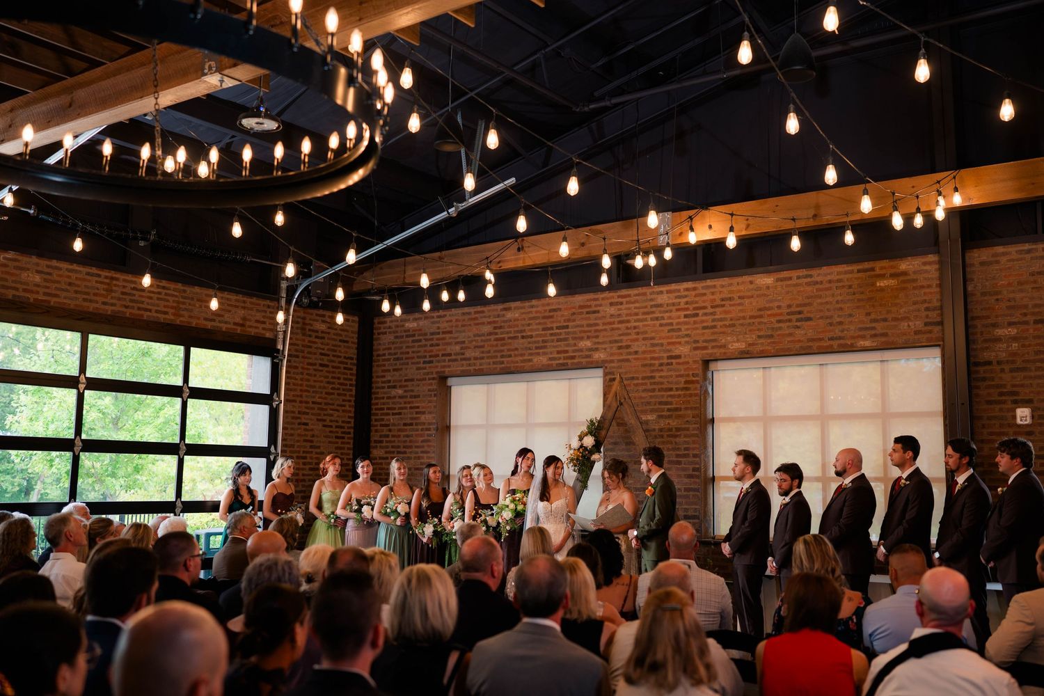 Full wedding party stands at the altar during ceremony in a rustic industrial brick venue with string lights.