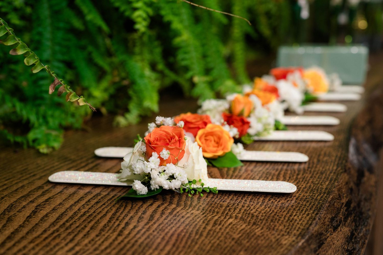 Row of orange and white floral boutonnieres on wooden sticks displayed on rustic table with ferns.