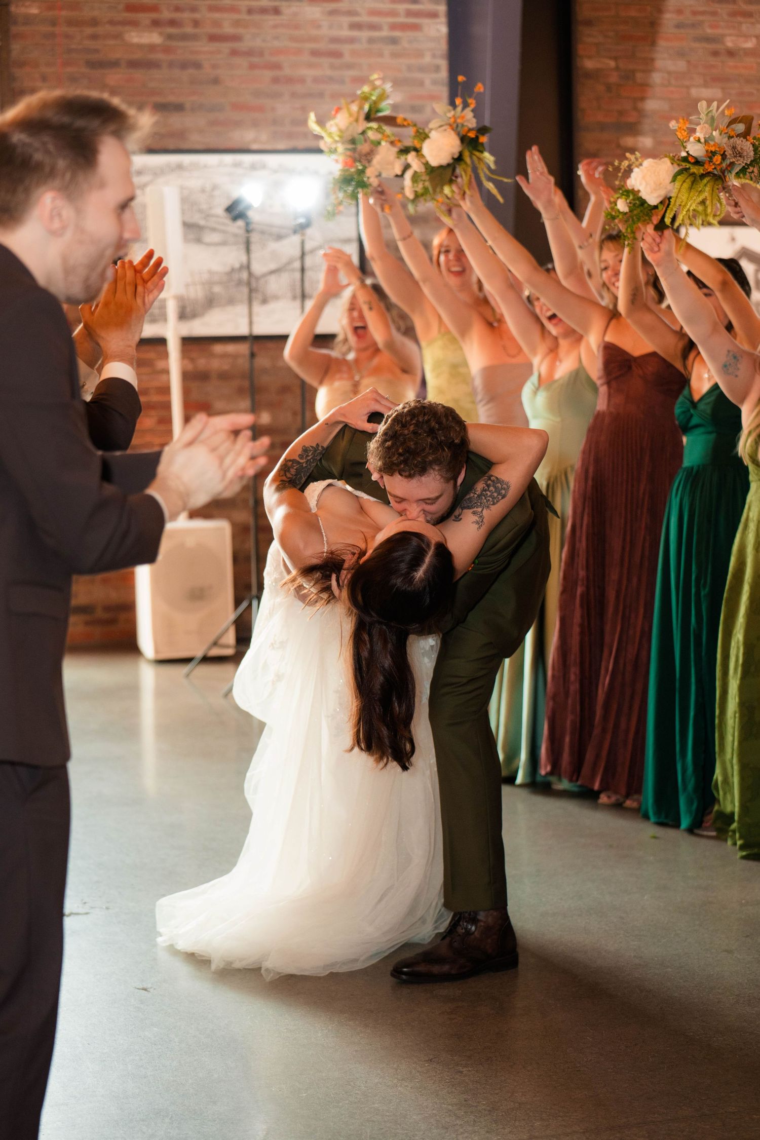 Newlyweds share romantic dip kiss surrounded by cheering wedding party in colorful gowns.