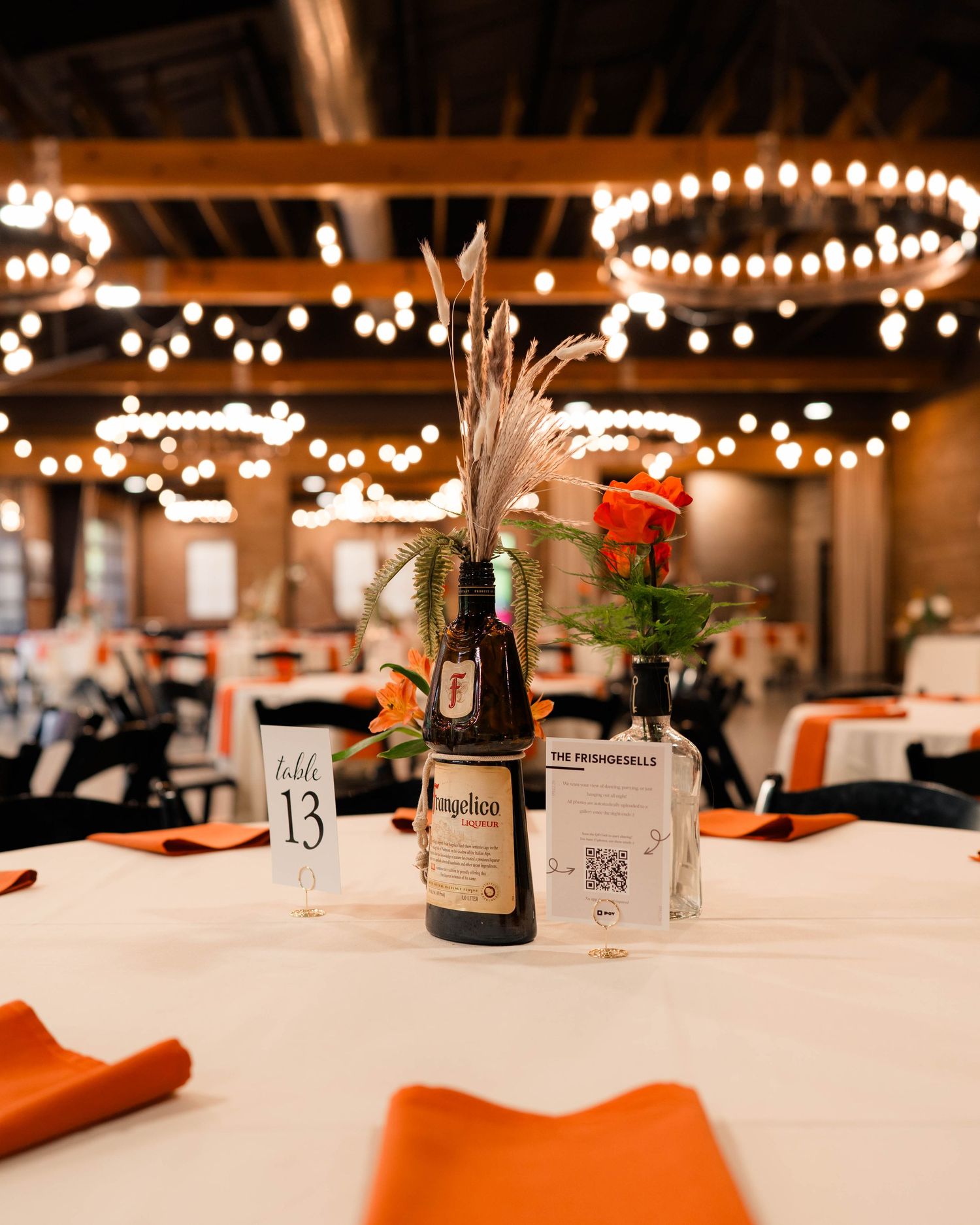 Elegant wedding reception table centerpiece featuring a Frangelico bottle, orange flowers, and string lights above.