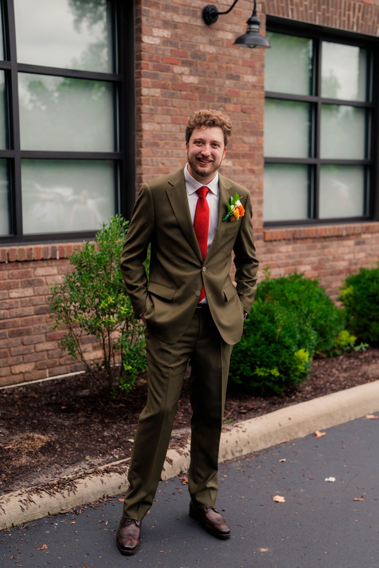 Smiling groom in olive green suit with red tie and floral boutonniere poses outside a brick building.