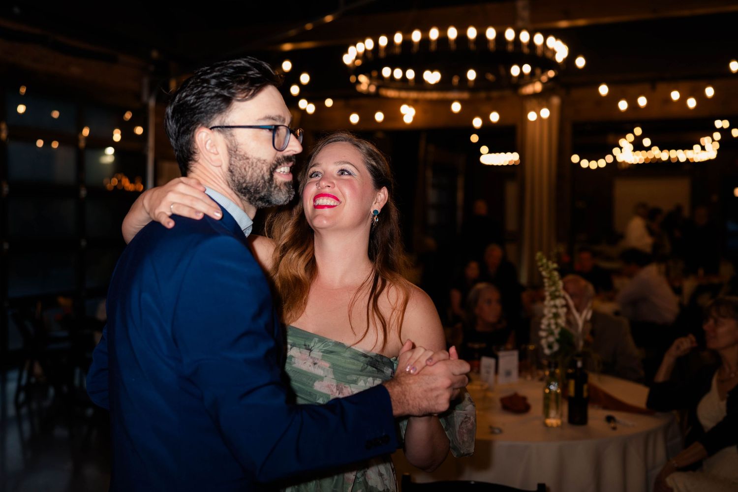 Couple sharing a dance and laughing together under chandelier lights at a wedding reception.