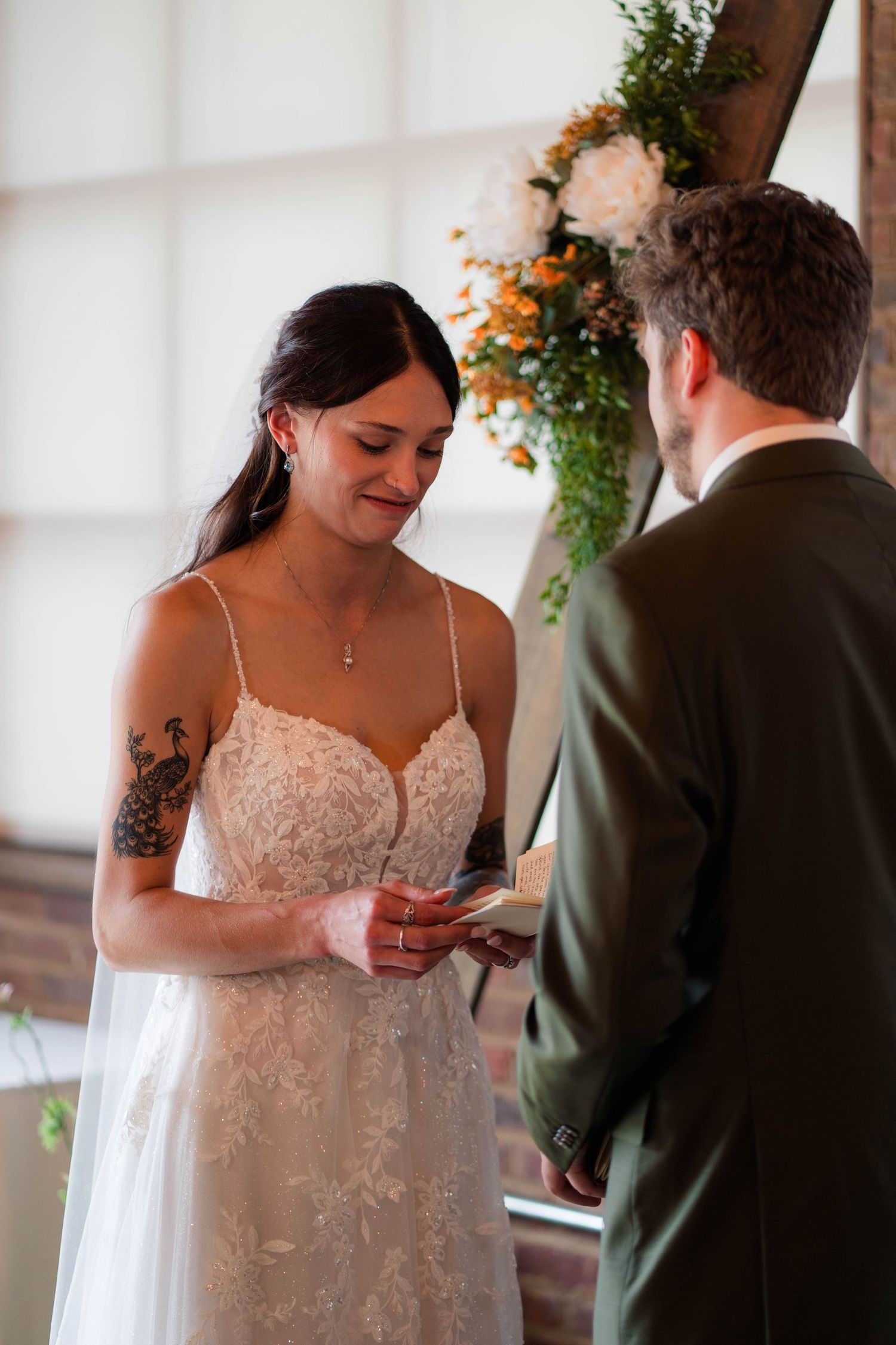 Bride with tattoos smiles while exchanging rings with groom during intimate indoor wedding ceremony.