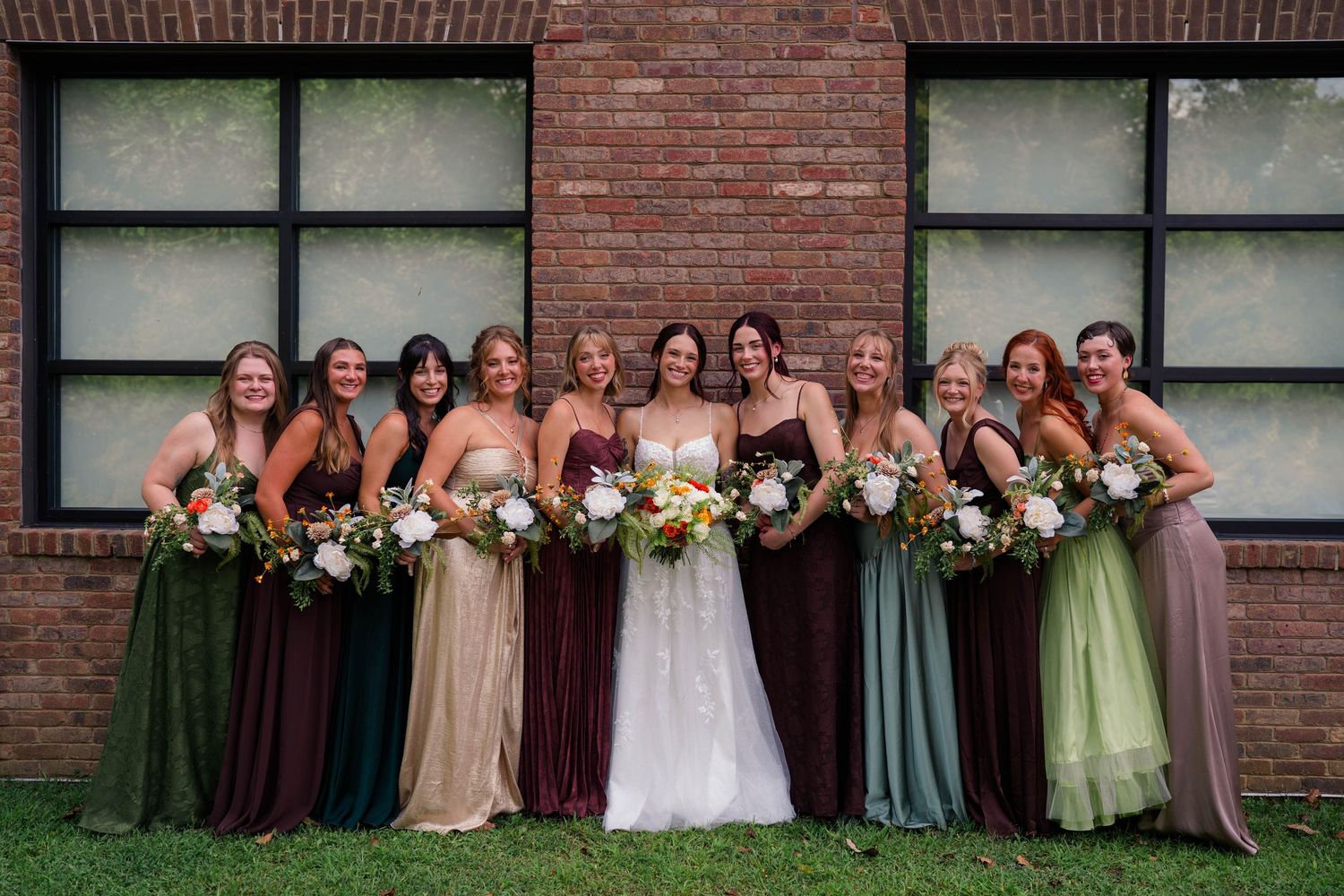 Bride poses with ten bridesmaids in mismatched earth-tone dresses holding bouquets outside brick building.