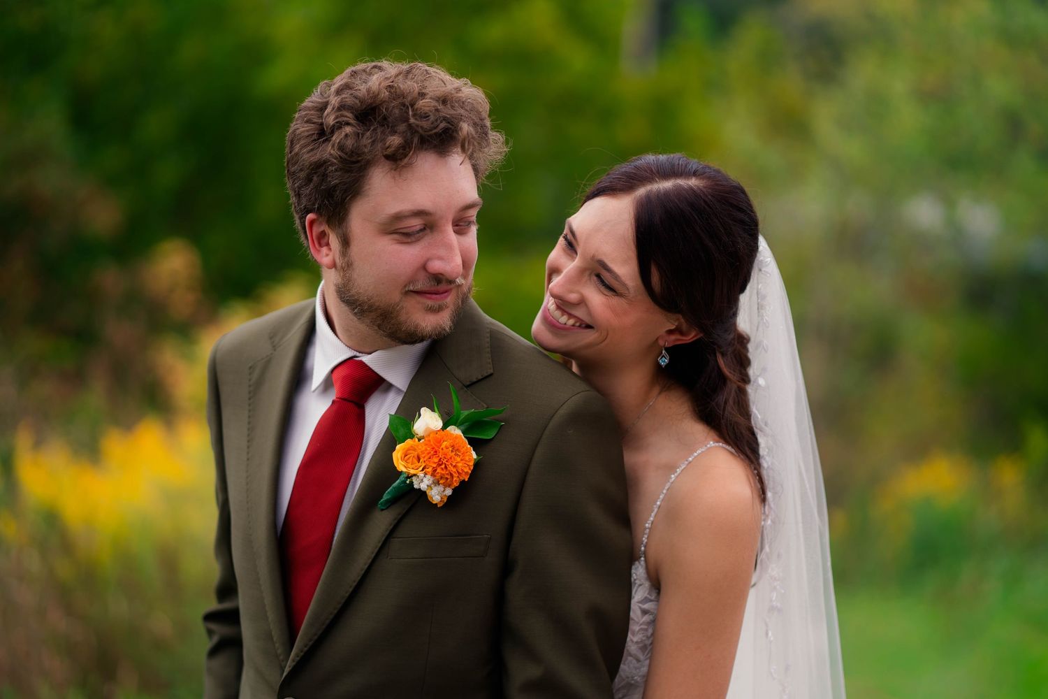 Bride and groom sharing a romantic outdoor moment, groom in olive suit with red tie and orange boutonniere.