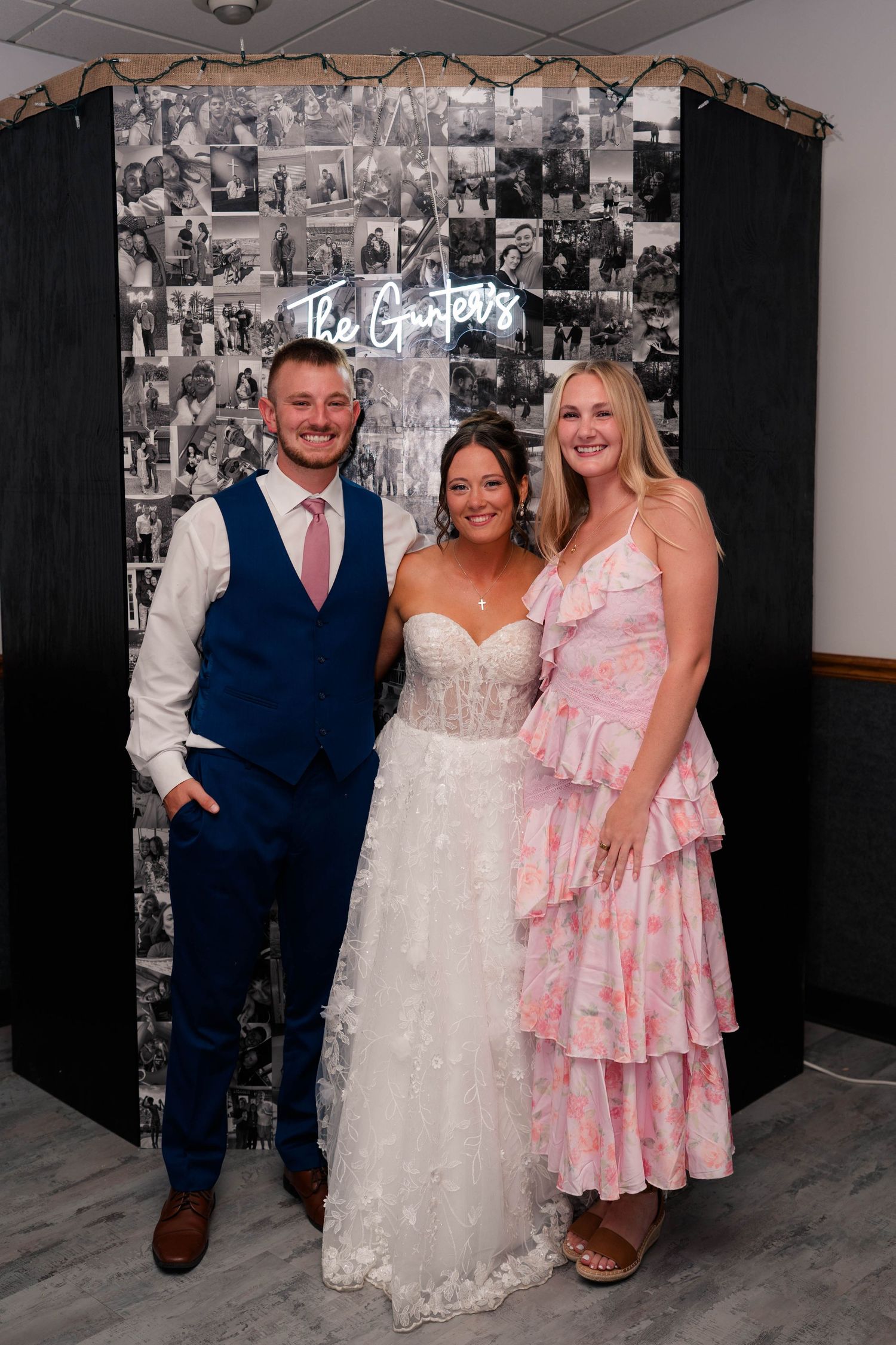 Bride in white gown poses with groom in blue suit and woman in pink dress at wedding reception photo wall.