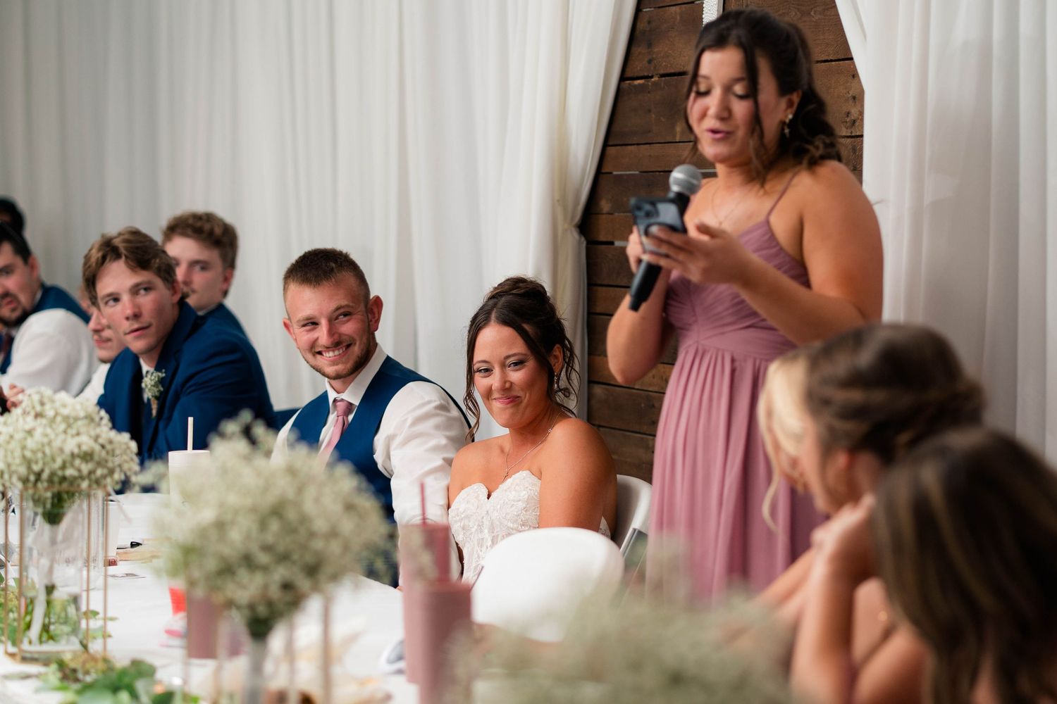 Bridesmaid in pink dress gives a heartfelt speech at wedding reception while bride and groom smile at the head table.