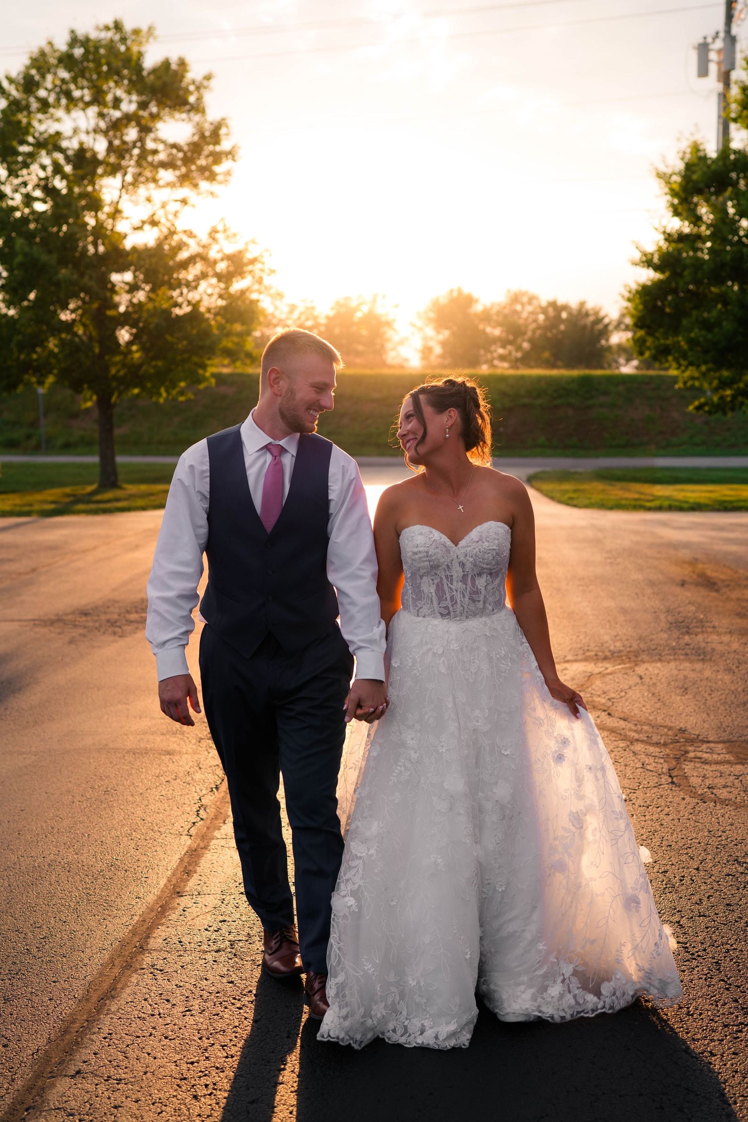 Bride and groom walk hand in hand down a road bathed in golden sunset light.
