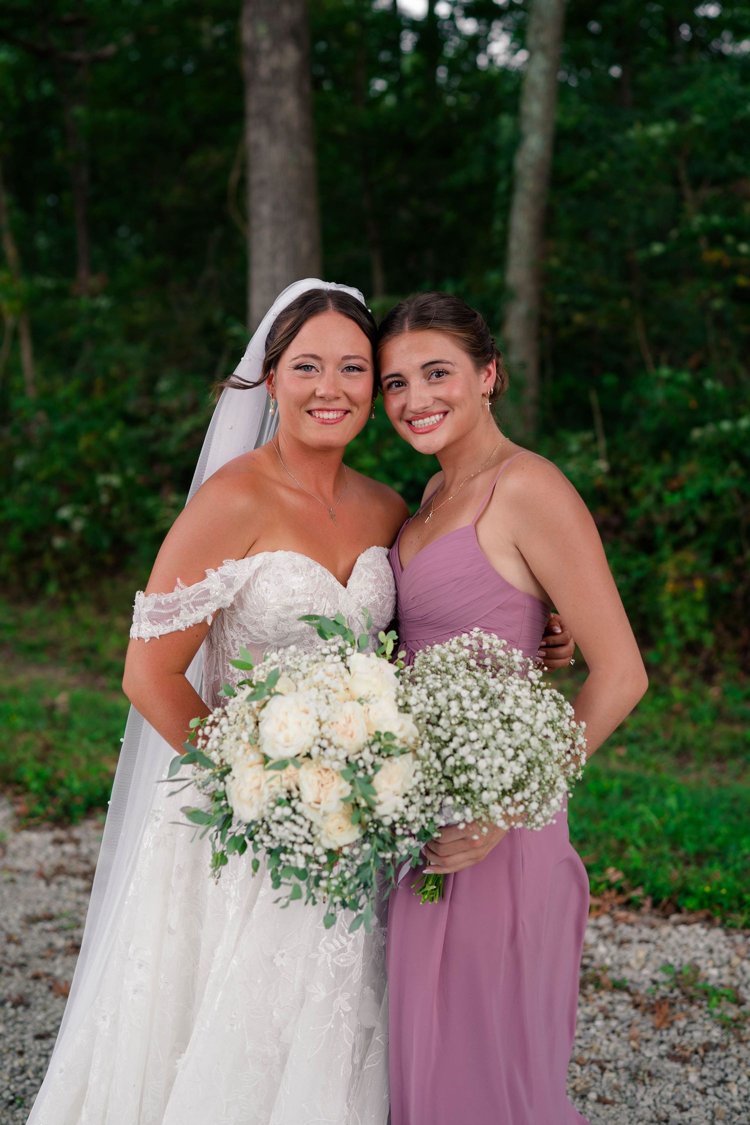 Smiling bride in off-shoulder lace gown poses with bridesmaid in mauve dress holding white floral bouquets outdoors.