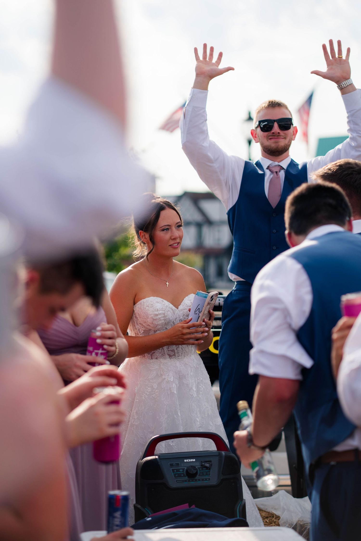 Bride and groom celebrating outdoors at wedding reception, groom raising hands wearing sunglasses.