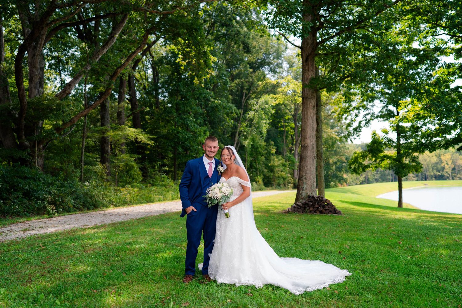 Bride and groom pose together on a green lawn surrounded by tall trees near a pond.
