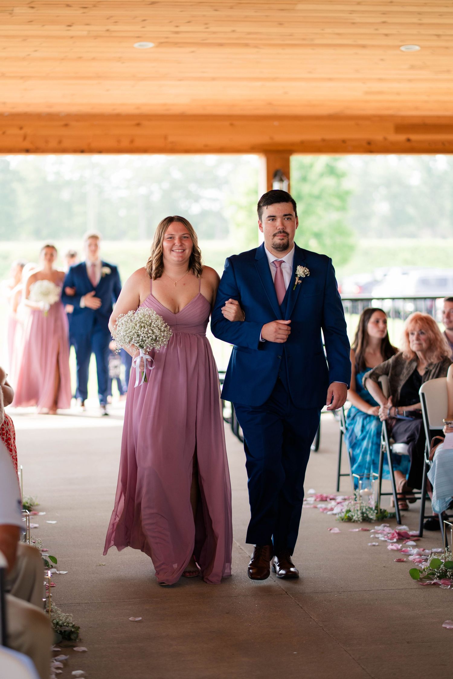 Bridesmaid in dusty rose gown walks down the aisle with groomsman in navy suit at wedding.