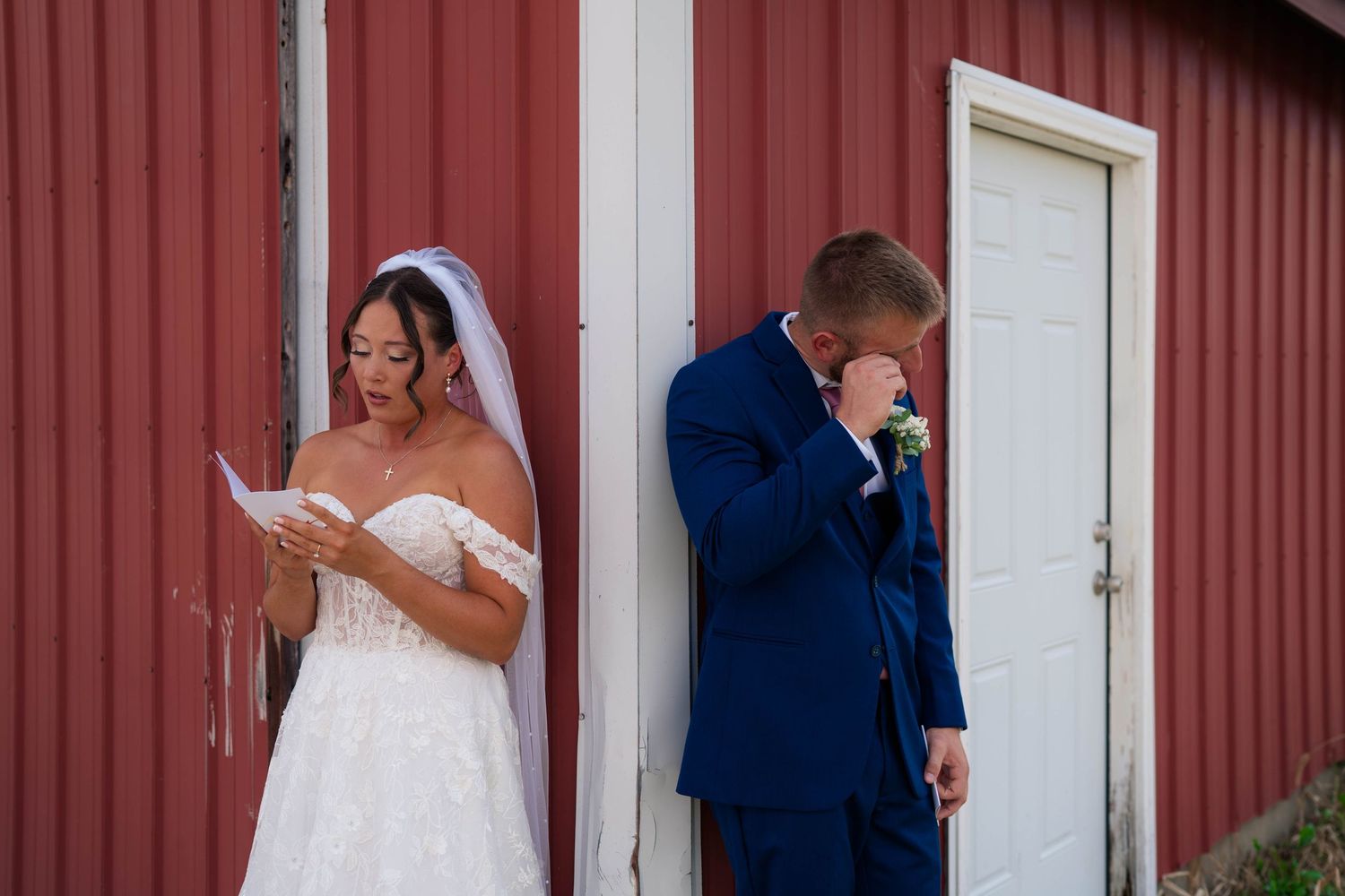 Emotional bride and groom exchange private vows outside red barn, groom wiping tears of joy.