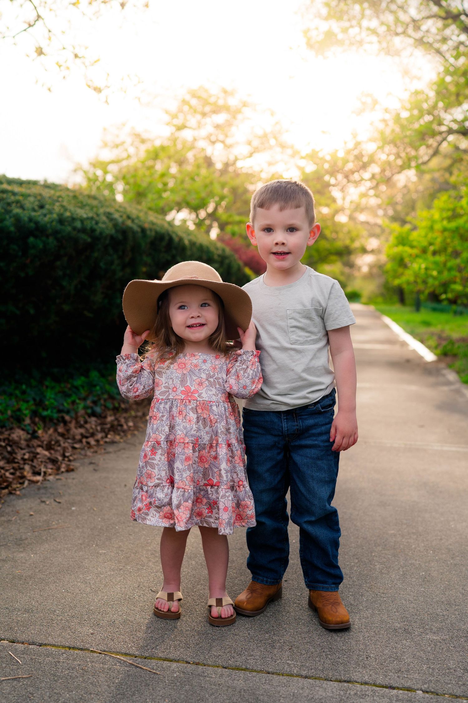Two young siblings pose on a sunny path, girl in floral dress and sun hat, boy in jeans and boots.