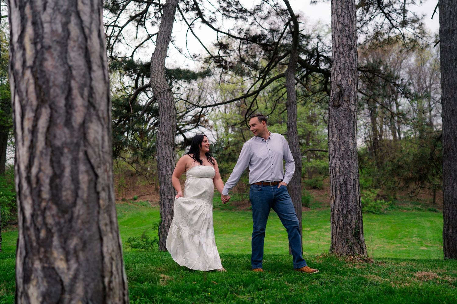 Couple holds hands smiling among tall trees on green lawn in romantic outdoor engagement photo session.