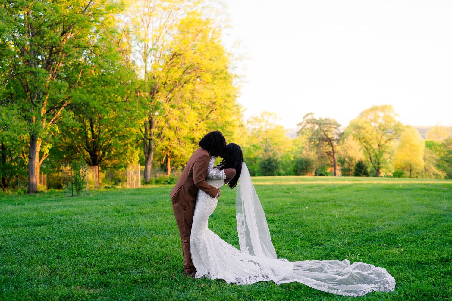 A bride and groom share a romantic kiss at golden hour in a lush green park, her long veil trailing behind.