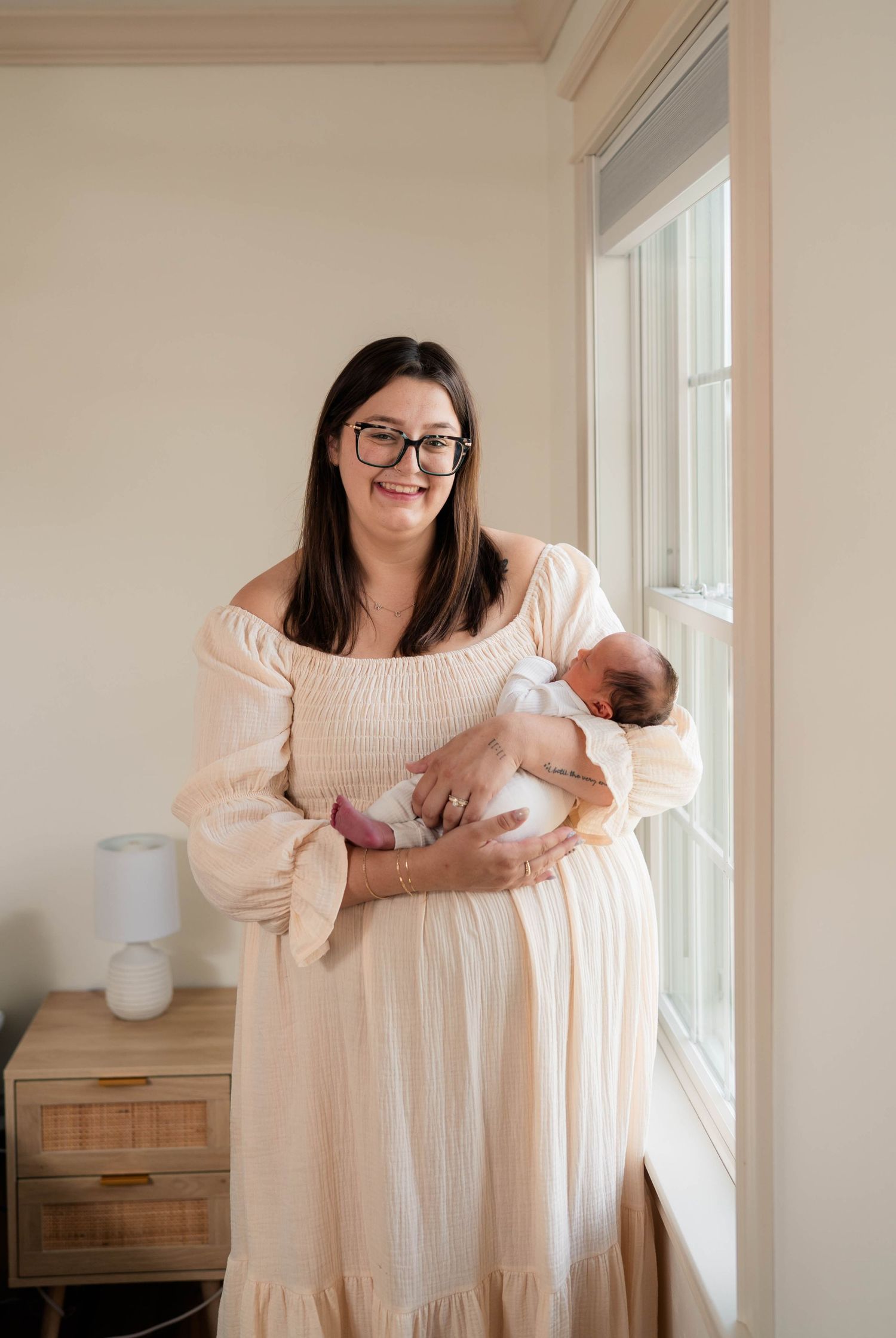 Smiling mother in cream dress holding newborn baby by sunlit window.