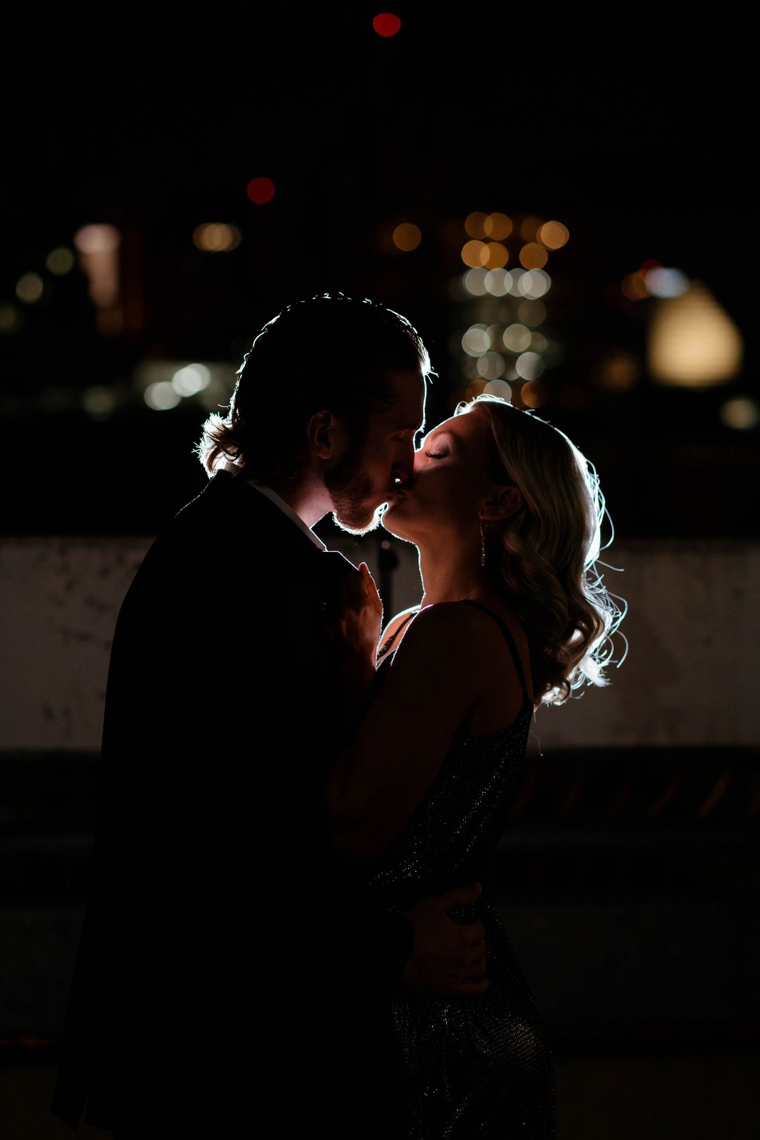 Silhouette of a couple kissing against a dark night sky with colorful bokeh city lights in the background.