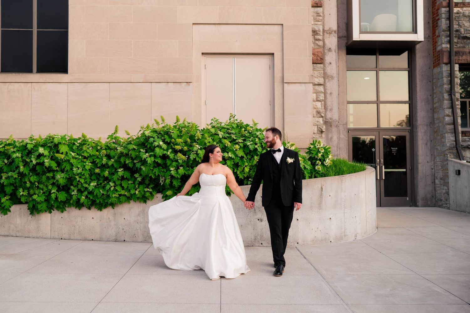 Bride twirls her white satin gown while walking with groom past a green hedge wall outside a stone venue.