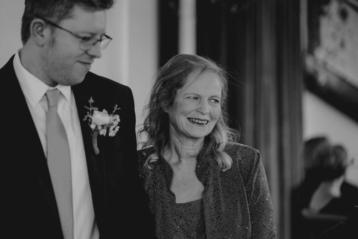 Black and white photo of smiling mother and groom with boutonniere at wedding ceremony.