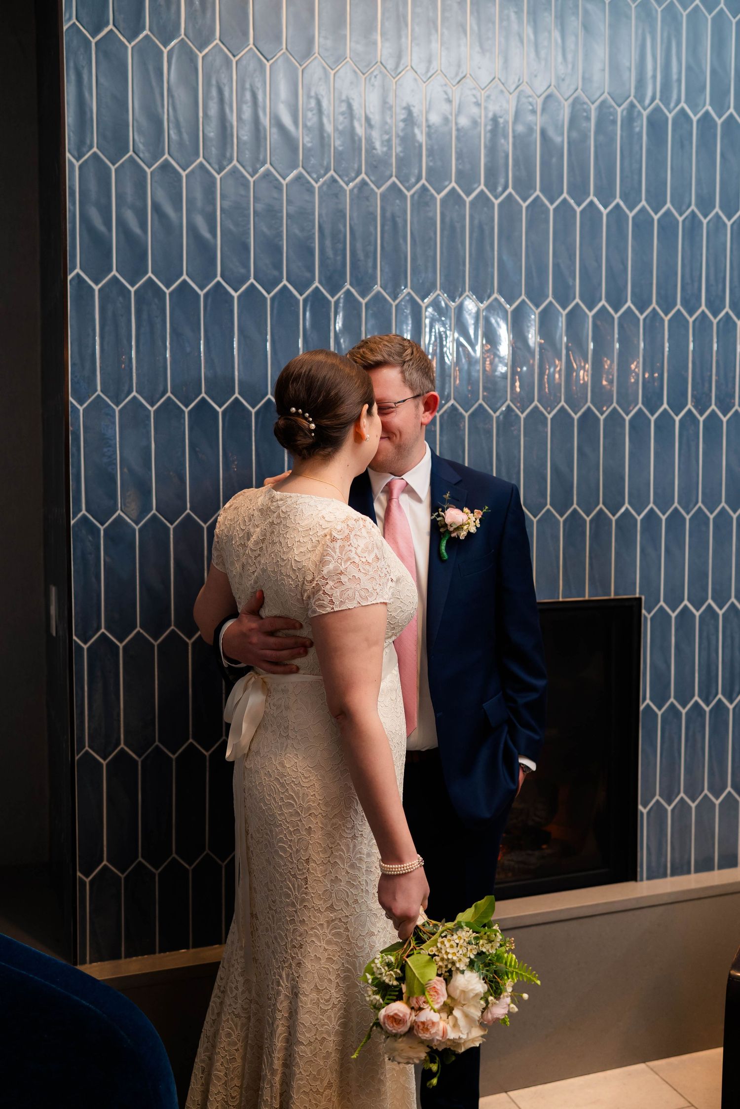 Bride and groom sharing an intimate moment by a blue tile fireplace, bride holding a pastel wedding bouquet.