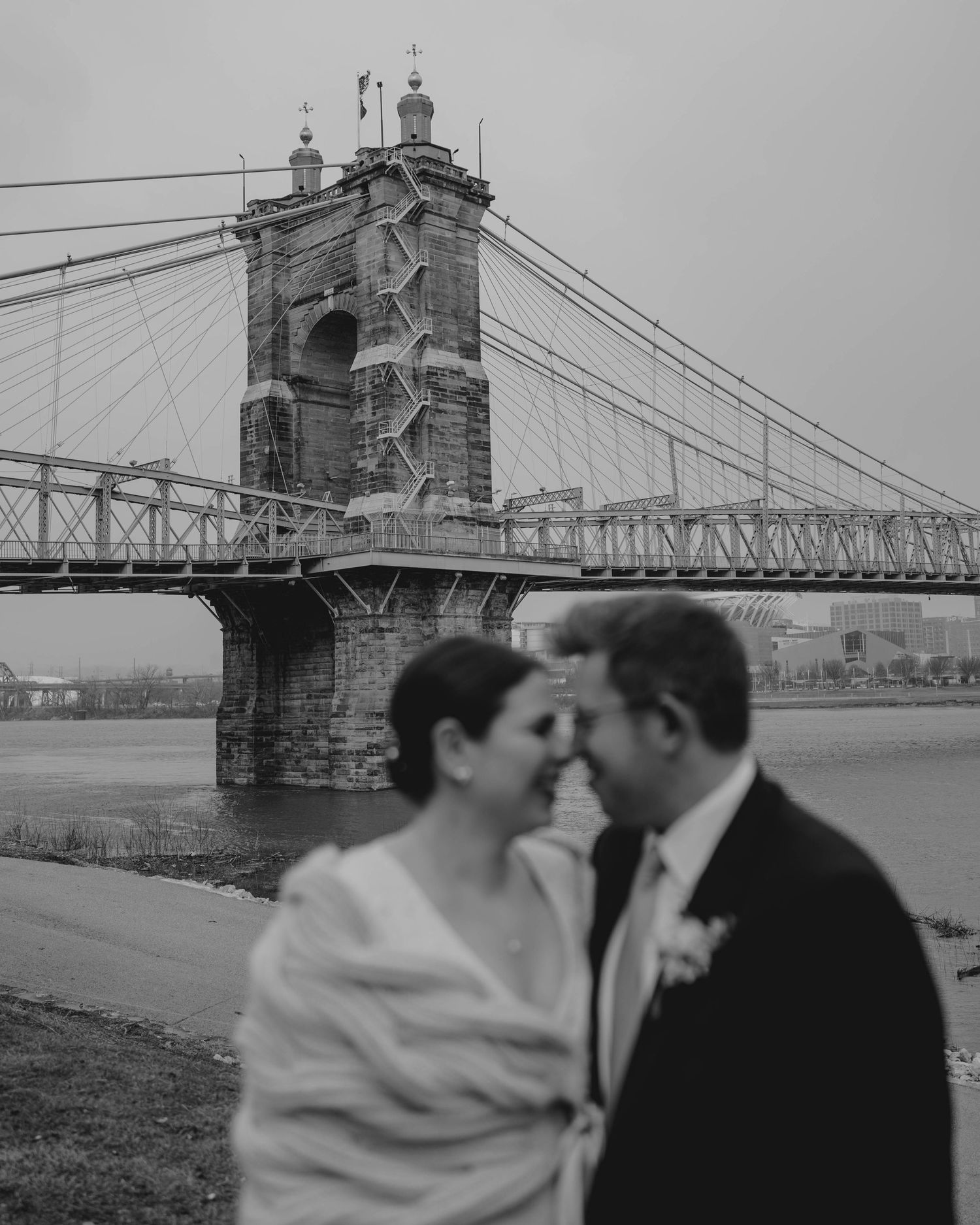 Artistic black and white photo of couple with Roebling Bridge tower dramatically framed behind them.