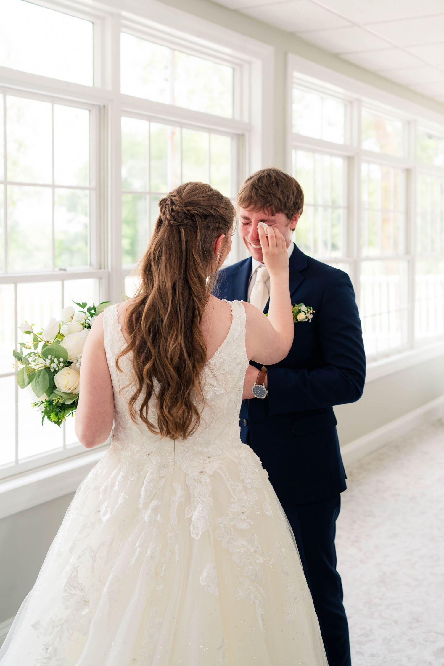 Emotional groom wipes tears seeing bride for first time during wedding day first look indoors.