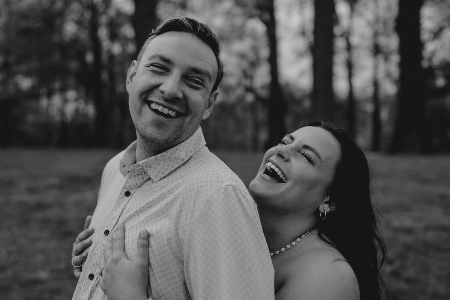 Black and white photo of a laughing couple embracing outdoors with trees in the background.