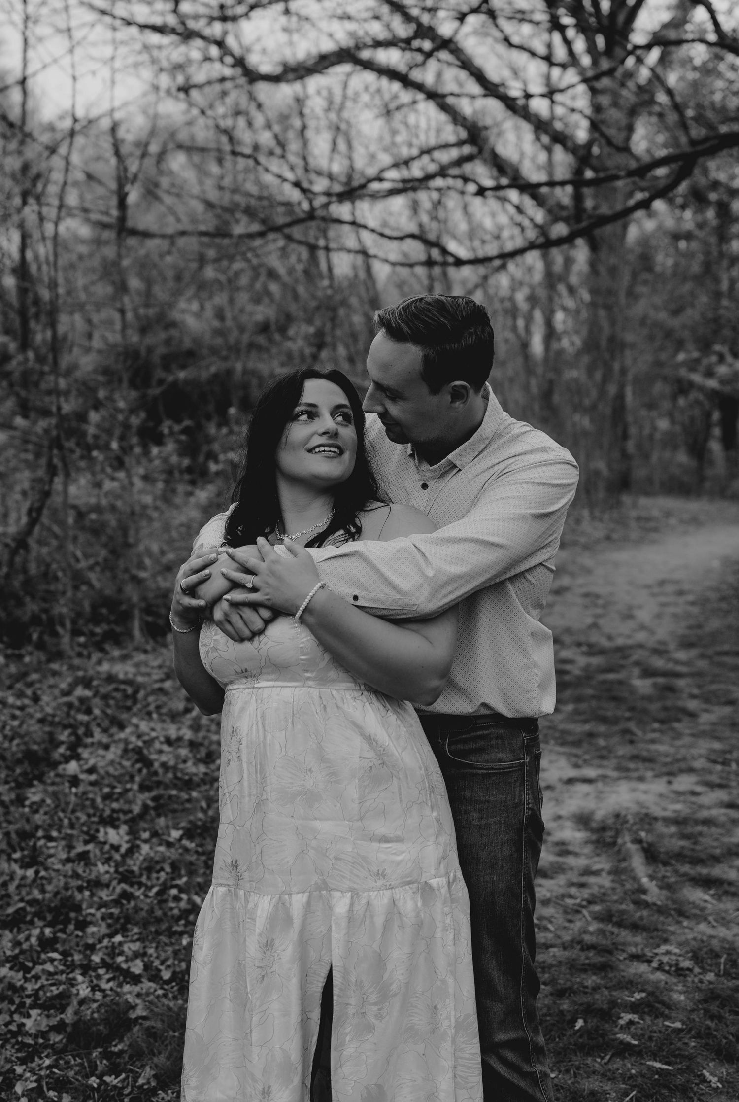 Black and white romantic portrait of couple embracing on woodland path, woman in white dress, man holding her tenderly.