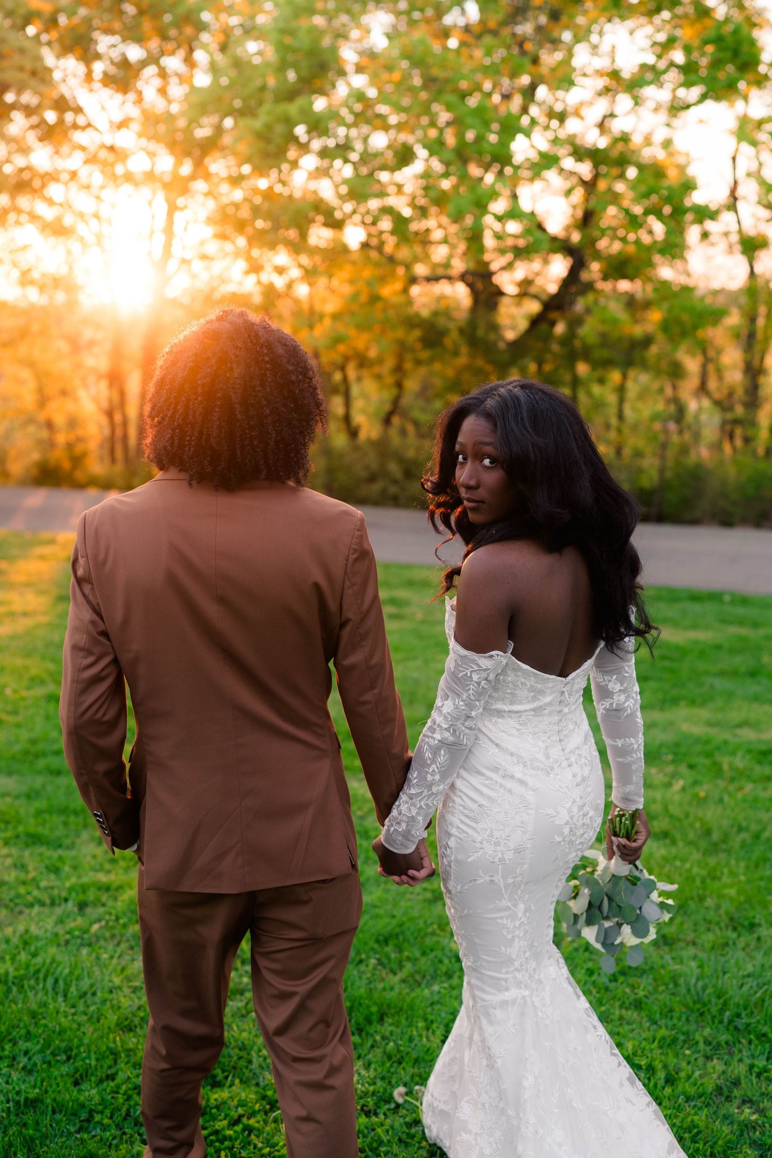 Bride in white lace gown holds bouquet, glancing back at groom in brown suit during golden hour outdoor wedding.