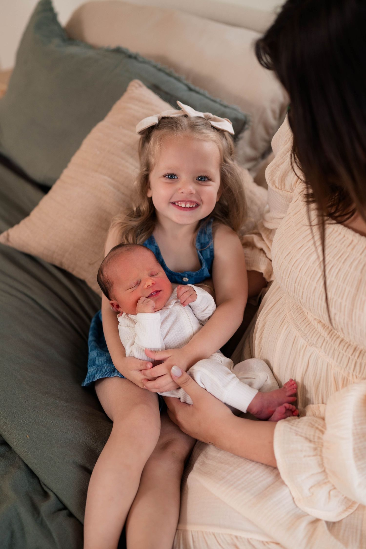 Smiling toddler girl with bow holds newborn baby while sitting on bed with mother in white dress.