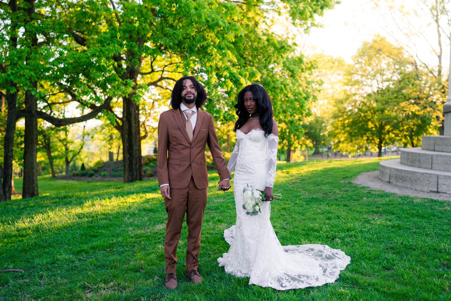 Bride in lace gown and groom in brown suit hold hands in sunlit park with green trees.