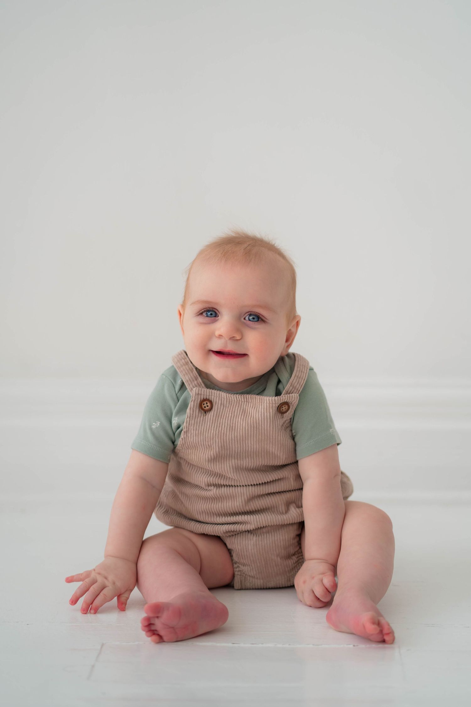 Smiling baby with blue eyes sitting on white floor wearing tan overalls and green shirt.