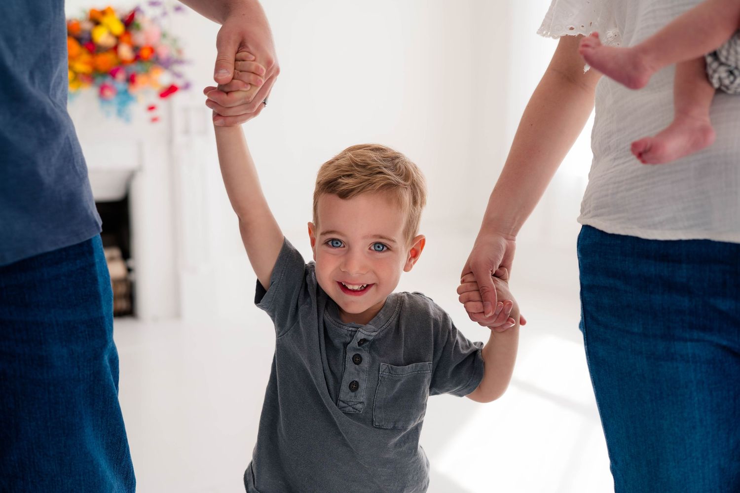 A happy toddler boy holds hands with his parents, smiling brightly in a bright white room.