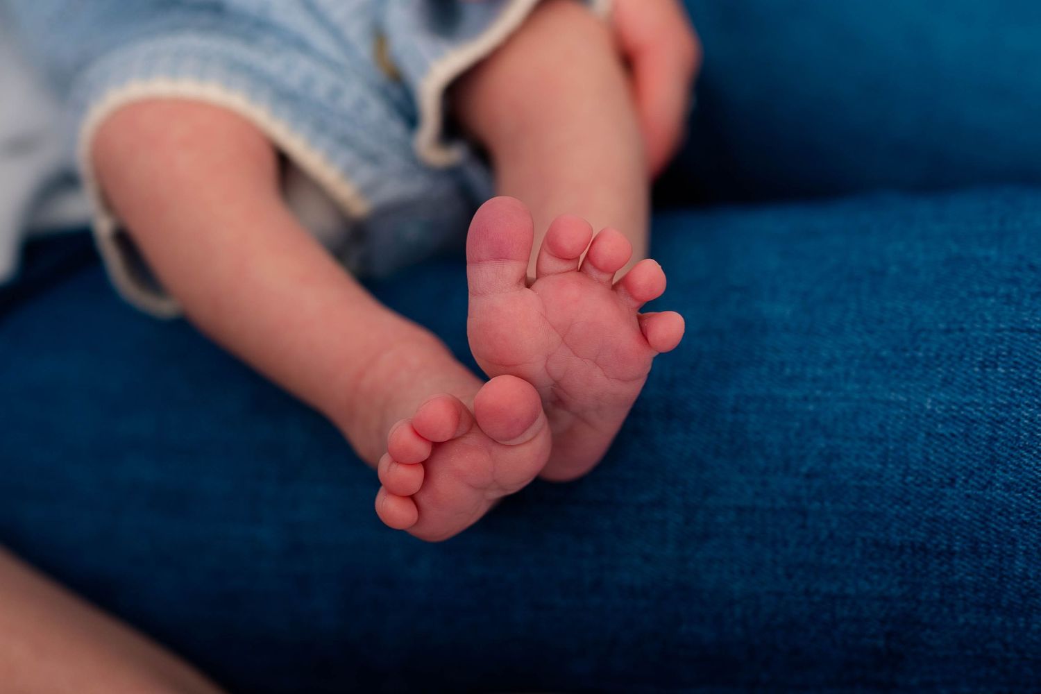 Tiny newborn baby feet rest on a blue denim surface in a close-up detail shot.