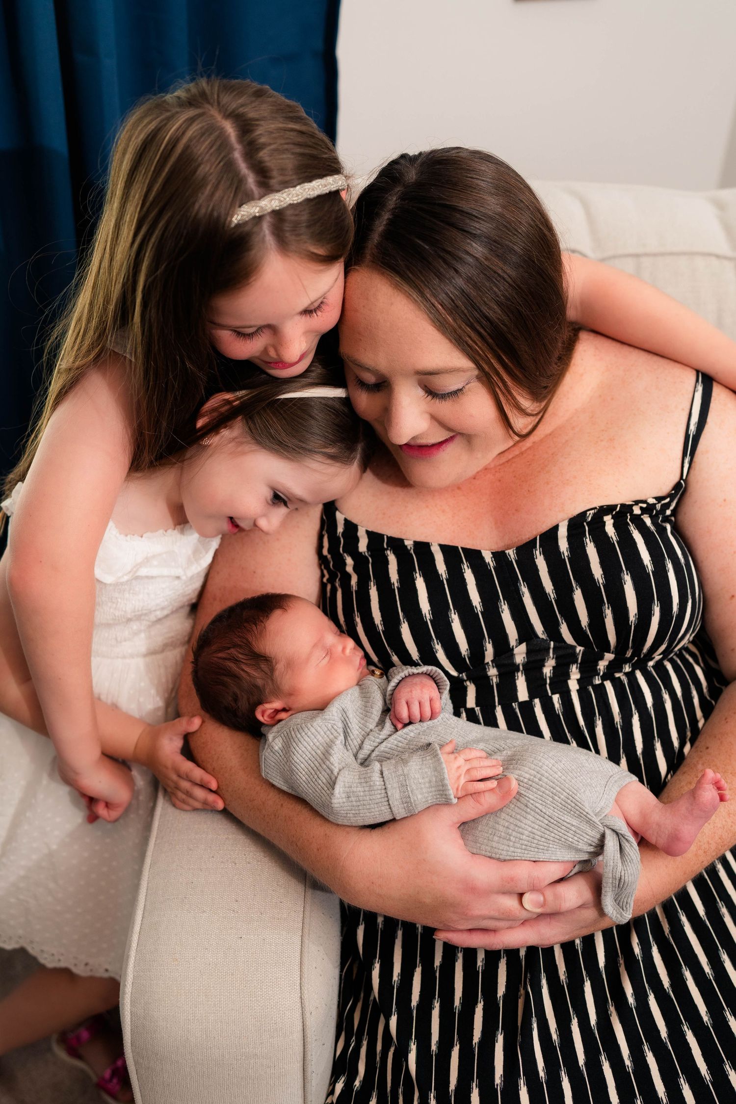 A mother in a striped dress holds her newborn baby while two young daughters lean in to admire their new sibling.