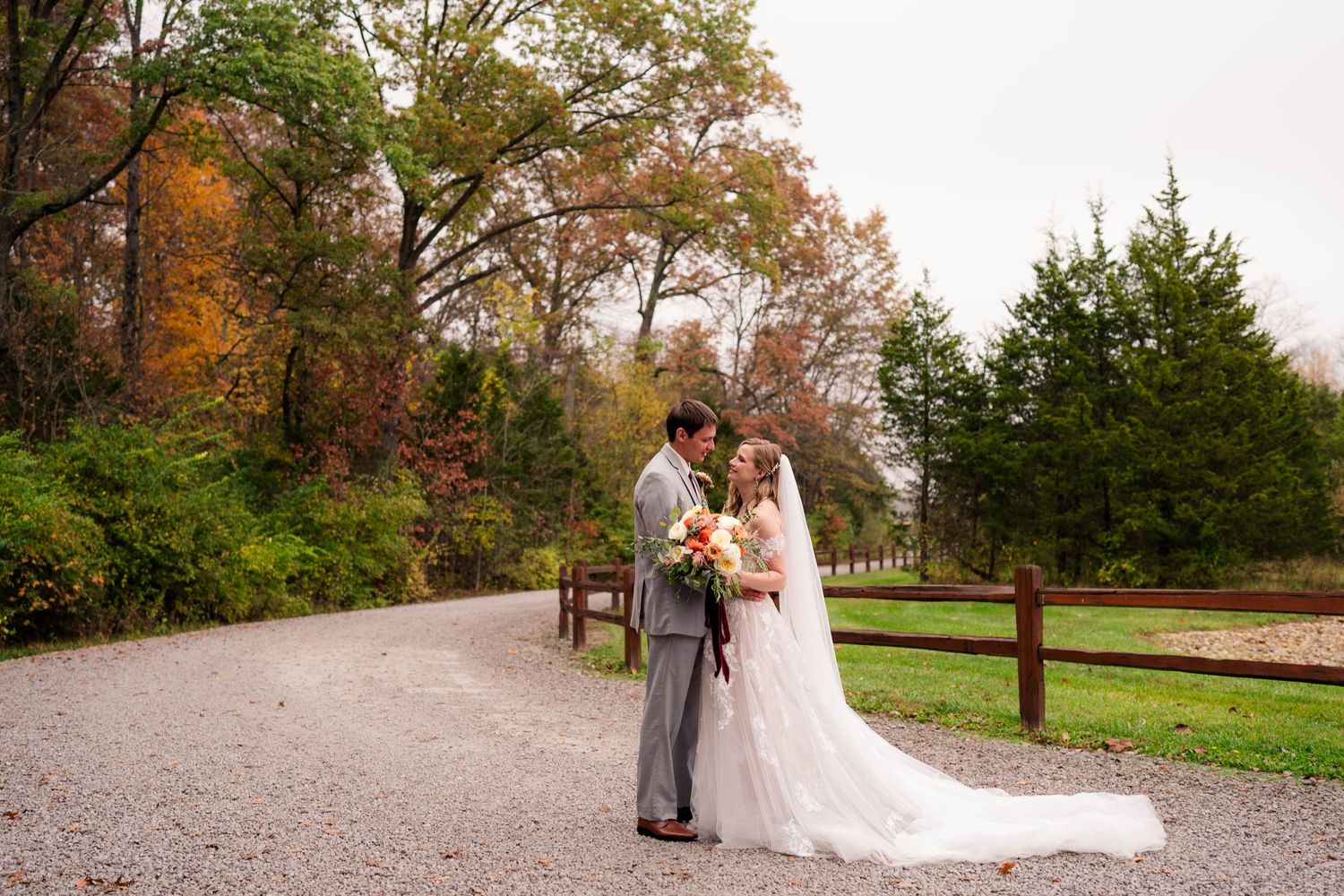 Bride and groom share a romantic moment on a gravel path surrounded by colorful autumn foliage at an outdoor wedding.