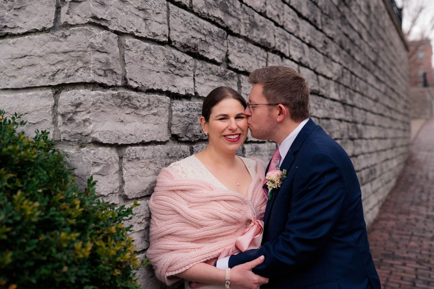 Groom in navy suit kisses bride on the cheek as she smiles against a rustic stone wall on their wedding day.