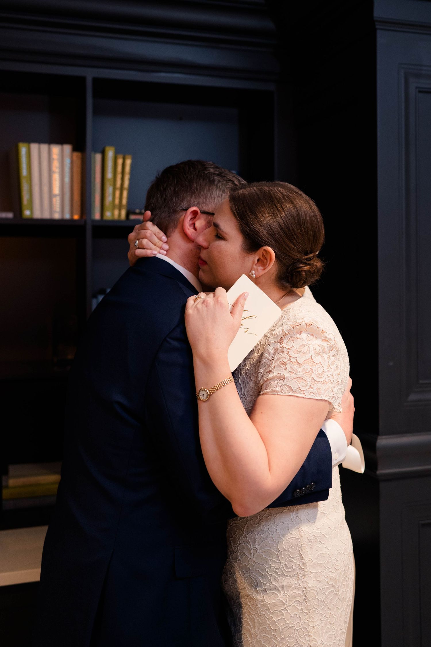 Bride and groom share a tender first kiss in a dark library setting on their wedding day.